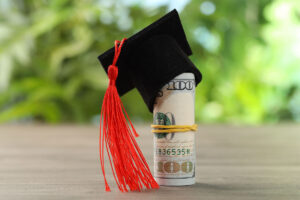 A rolled-up U.S. hundred dollar bill secured with a rubber band, wearing a black graduation cap with a red tassel, sits on a wooden surface with a blurred green background.