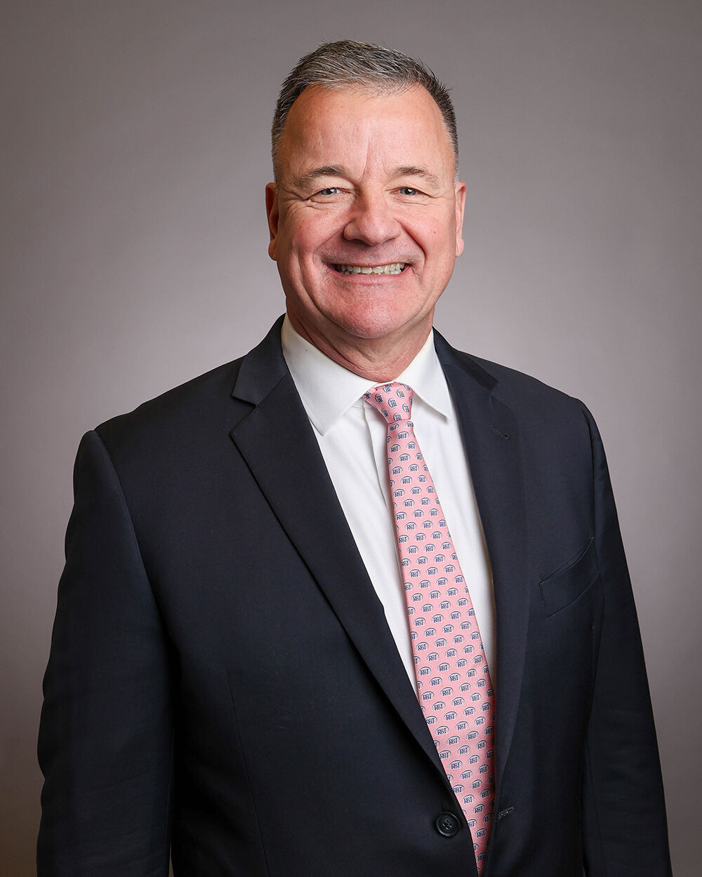 A middle-aged man with short gray hair is smiling and wearing a dark suit, white shirt, and a light pink patterned tie, standing against a neutral gray background.