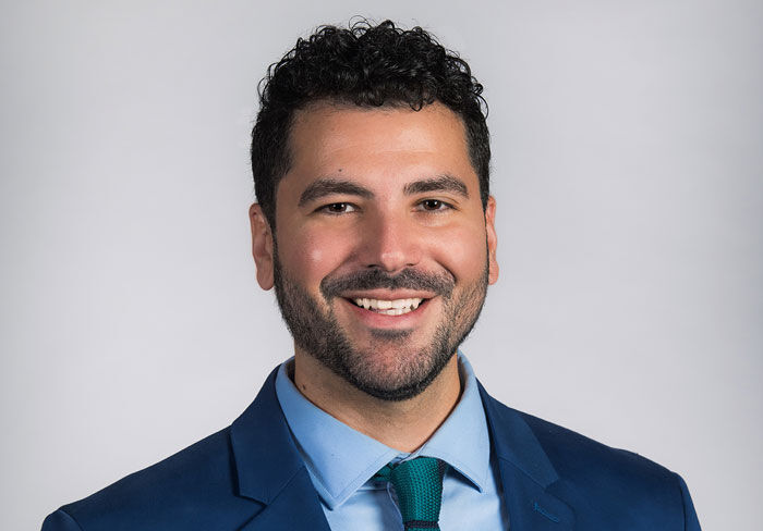 A man with short, curly dark hair and a beard, wearing a blue suit, light blue shirt, and green tie, smiles against a plain light gray background.