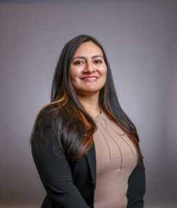 A woman with long dark hair wearing a beige top and a black blazer is smiling and posing for a professional portrait against a plain gray background.