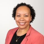 A woman with short curly hair smiles at the camera. She is wearing a coral blazer over a black top and a gold necklace with a small pendant, against a plain light background.