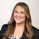 A woman with long, wavy blonde hair smiles at the camera. She is wearing a dark blazer over a patterned top and is positioned in front of a plain, light background.