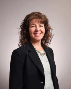 A woman with curly brown hair wearing a black blazer, a light blue lace top, and a statement necklace, smiling at the camera against a plain light background.