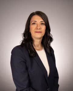 A woman with long dark hair wearing a navy blazer over a light top, smiling slightly, poses in front of a neutral gray background.