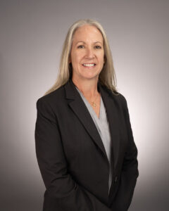 A woman with long gray hair wearing a black blazer and gray blouse smiles at the camera against a plain, light gray background.