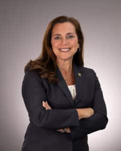 A woman with long brown hair in a dark blazer stands with her arms crossed, smiling confidently against a plain, light gray background.