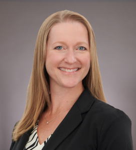 A woman with straight, shoulder-length blonde hair smiles at the camera. She is wearing a black blazer over a patterned blouse, with a neutral gray background.