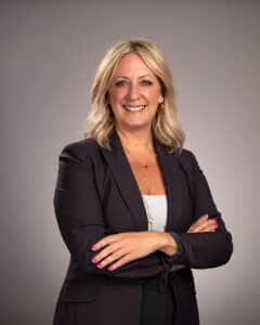 A woman with blonde hair, wearing a dark blazer over a light top, smiles confidently with her arms crossed in front of a plain, neutral background. She has pink nail polish and gold hoop earrings.