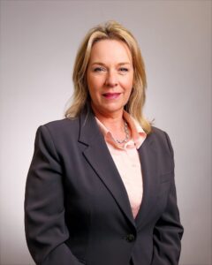 A woman with blonde hair wearing a dark blazer over a light pink blouse and a silver necklace, poses for a professional portrait against a neutral background.