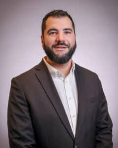 A man with short dark hair and a full beard is wearing a dark suit jacket over a white dress shirt, standing against a neutral light background and smiling slightly at the camera.