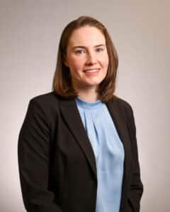 A woman with straight brown hair wearing a black blazer over a light blue blouse stands smiling in front of a plain, light-colored background.