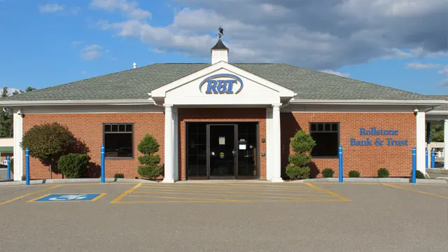 Parkhill A brick building with a sign reading "RBT" above the entrance is shown. The building has a green roof and white trim, with small trees and shrubs flanking the entrance. The words "Rollstone Bank & Trust" are visible on the right side of the building.