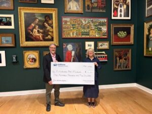 Two people stand in front of a green wall covered with framed artwork, holding a large ceremonial check for $1,020,000 from Baltimore Bank & Trust made out to the Fitzhugh Art Museum.
