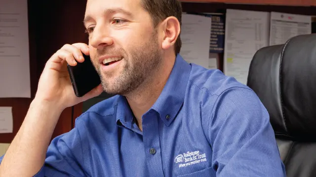 RBTman-Phone A man with a beard, wearing a blue shirt, smiles as he talks on a phone. He is seated in an office chair with documents pinned on the wall behind him. The shirt has an embroidered logo that reads "Bankers Bank of the West" above some smaller, unreadable text.