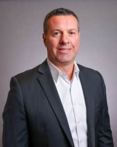 A man in a dark suit jacket and white dress shirt is standing and smiling at the camera against a plain, light gray background.