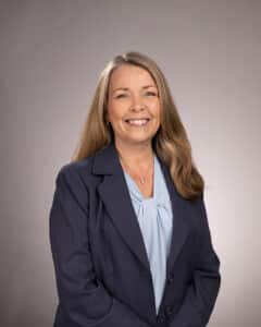 A woman with long, light brown hair smiles at the camera. She is wearing a dark pinstripe blazer over a light blue blouse, standing in front of a plain, light gray background.