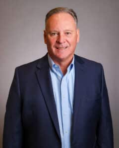 A middle-aged man with short, light brown hair wears a blue suit jacket over a light blue dress shirt, standing in front of a plain gray background and smiling at the camera.