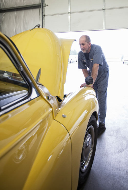 A mechanic in gray coveralls inspects the engine of a yellow vintage car with its hood open inside a garage.
