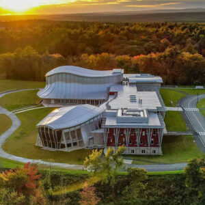 Aerial view of a modern, curved-roof building surrounded by trees and green lawns at sunset, with a winding road and scenic landscape in the background.
