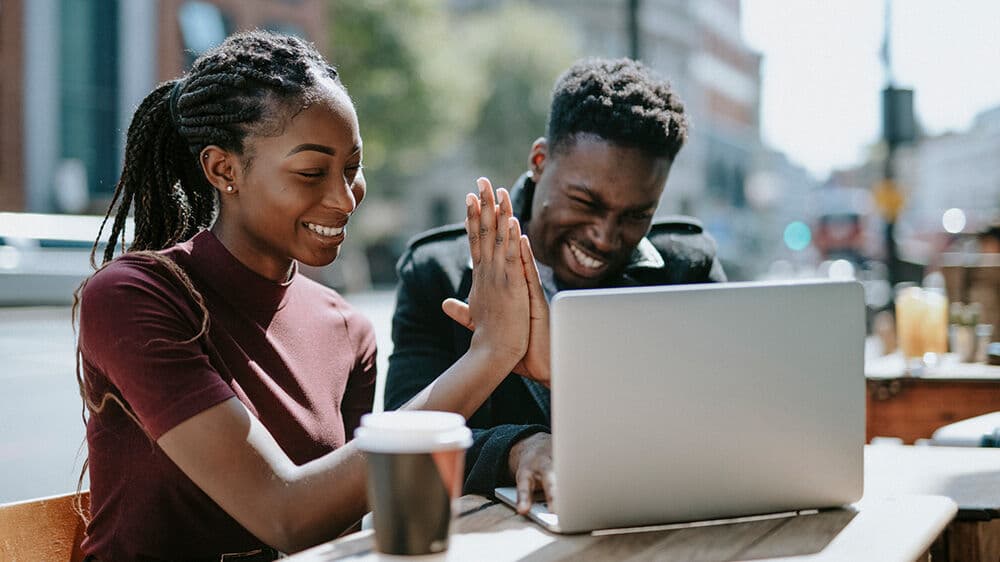 Two people sit at an outdoor table with a laptop, smiling and giving each other a high five. There is a coffee cup on the table, and city buildings are visible in the background.