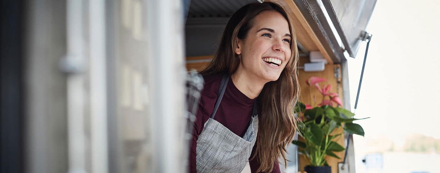 A smiling woman wearing an apron stands inside a food truck, looking outside. There is a green potted plant with pink flowers visible beside her, and natural light is coming in.