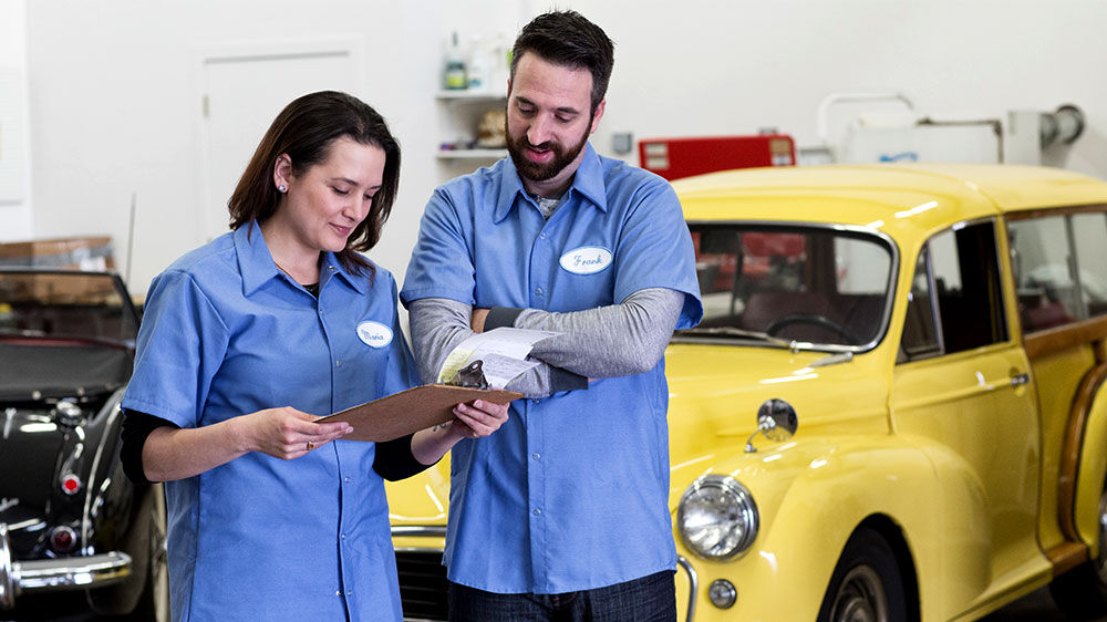 Two auto shop workers in blue uniforms review a clipboard together while standing in front of a vintage yellow car inside a garage.