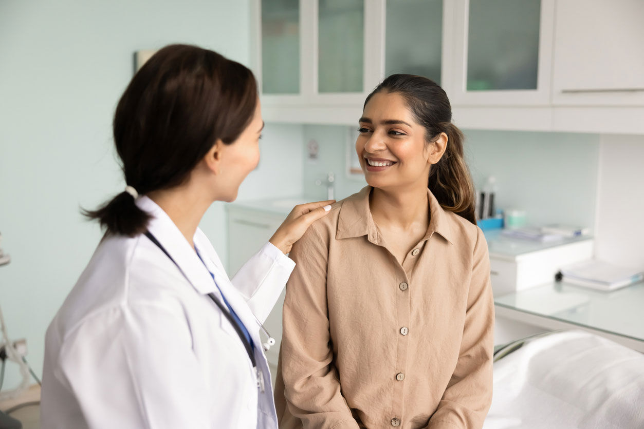 A female doctor wearing a white coat gently places her hand on a smiling woman’s shoulder as they sit and talk in a bright, modern medical office.