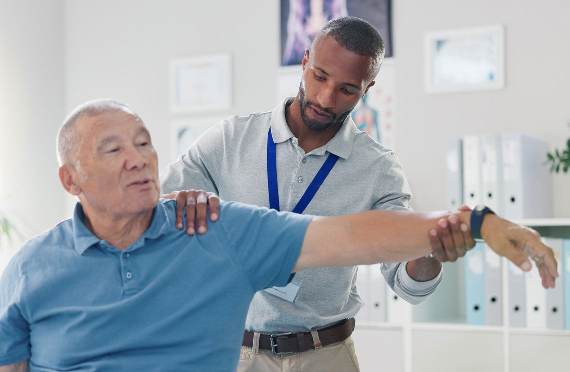 A physical therapist assists an older man in a blue shirt by gently stretching his extended arm during a rehabilitation session in a bright medical office.