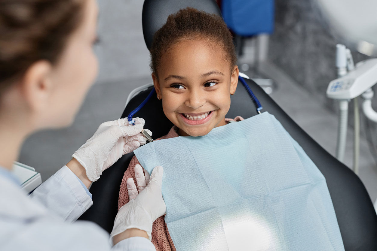 A young girl sits in a dentist’s chair smiling, wearing a blue dental bib. A dentist wearing gloves holds a dental tool and talks to her. The scene looks friendly and reassuring in a dental clinic.