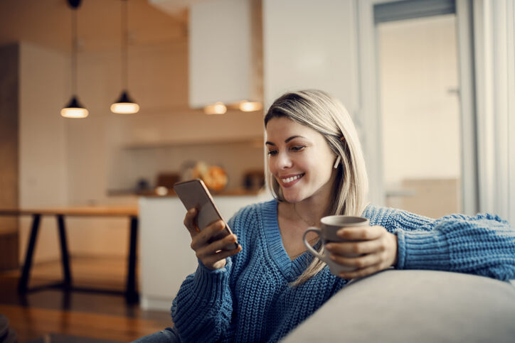 A woman in a blue sweater smiles while holding a smartphone and a mug of coffee.