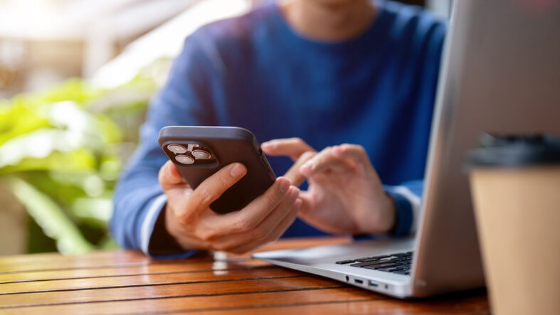 A person in a blue sweater using a smartphone while sitting at a table with a laptop and a coffee cup.