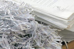 A pile of shredded paper strips sits next to a neatly stacked pile of intact documents on a desk.